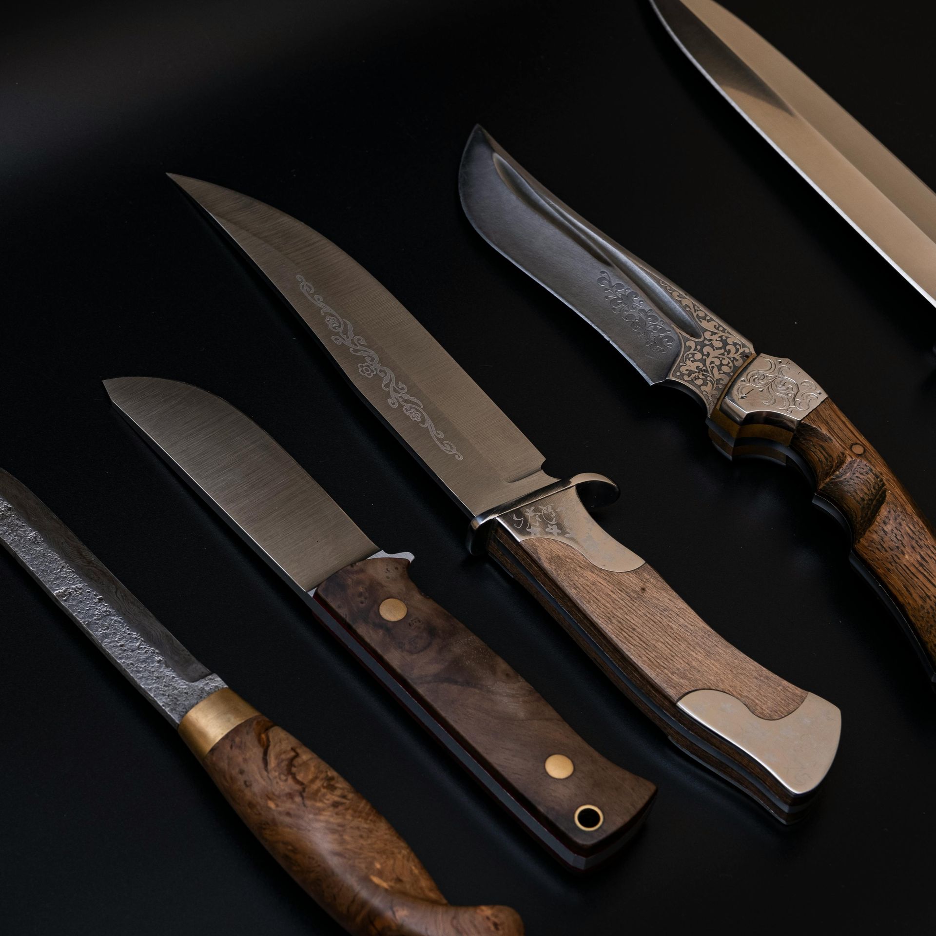 Four custom knives with ornate blades and textured wooden handles laid diagonally on a solid black surface.