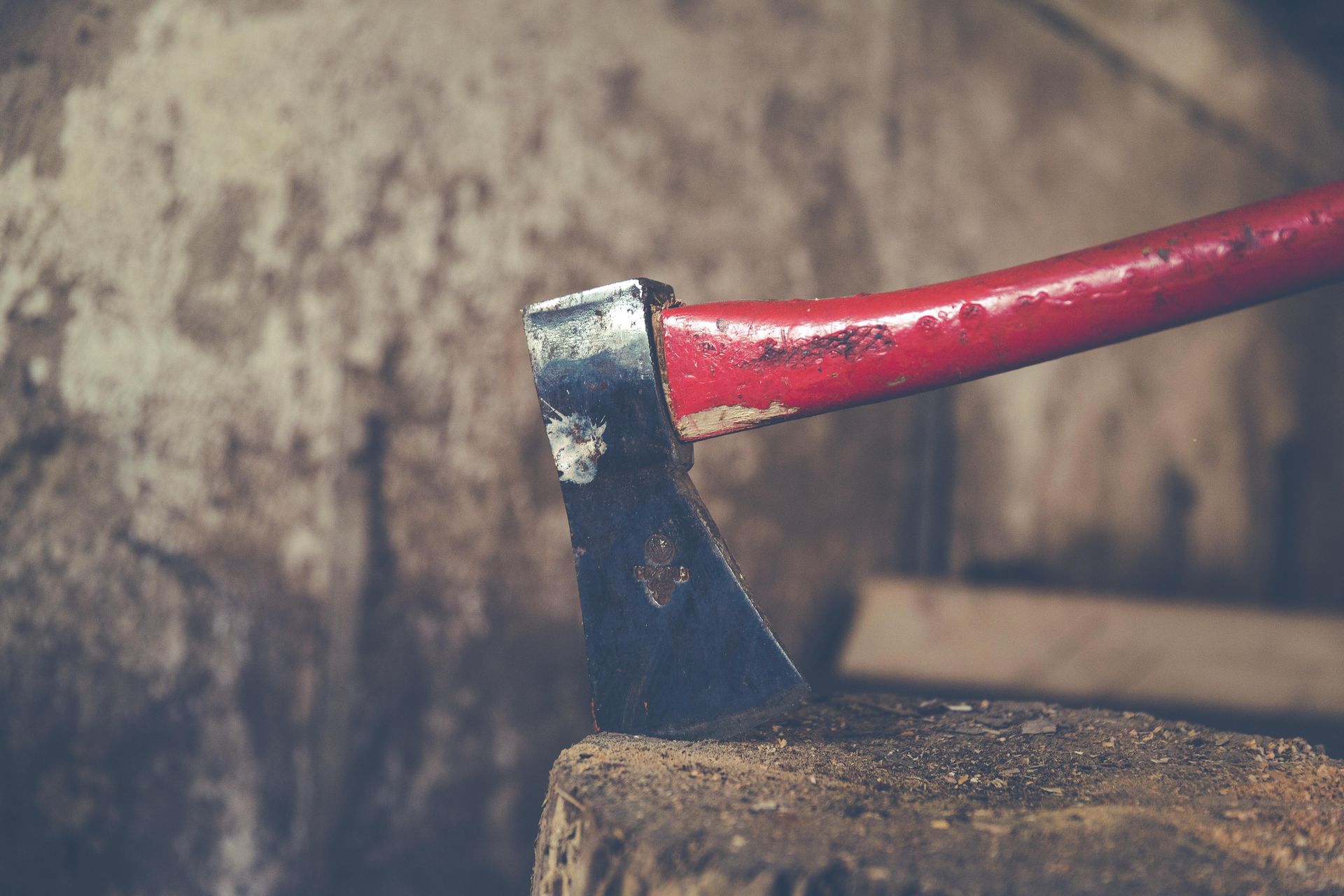 A rusty axe with a red handle rests on a wooden block in a dimly lit, textured space.