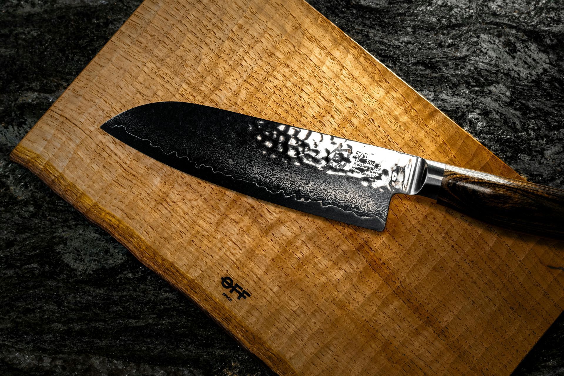 Japanese chef's knife on a wooden cutting board, resting on a dark, textured surface.