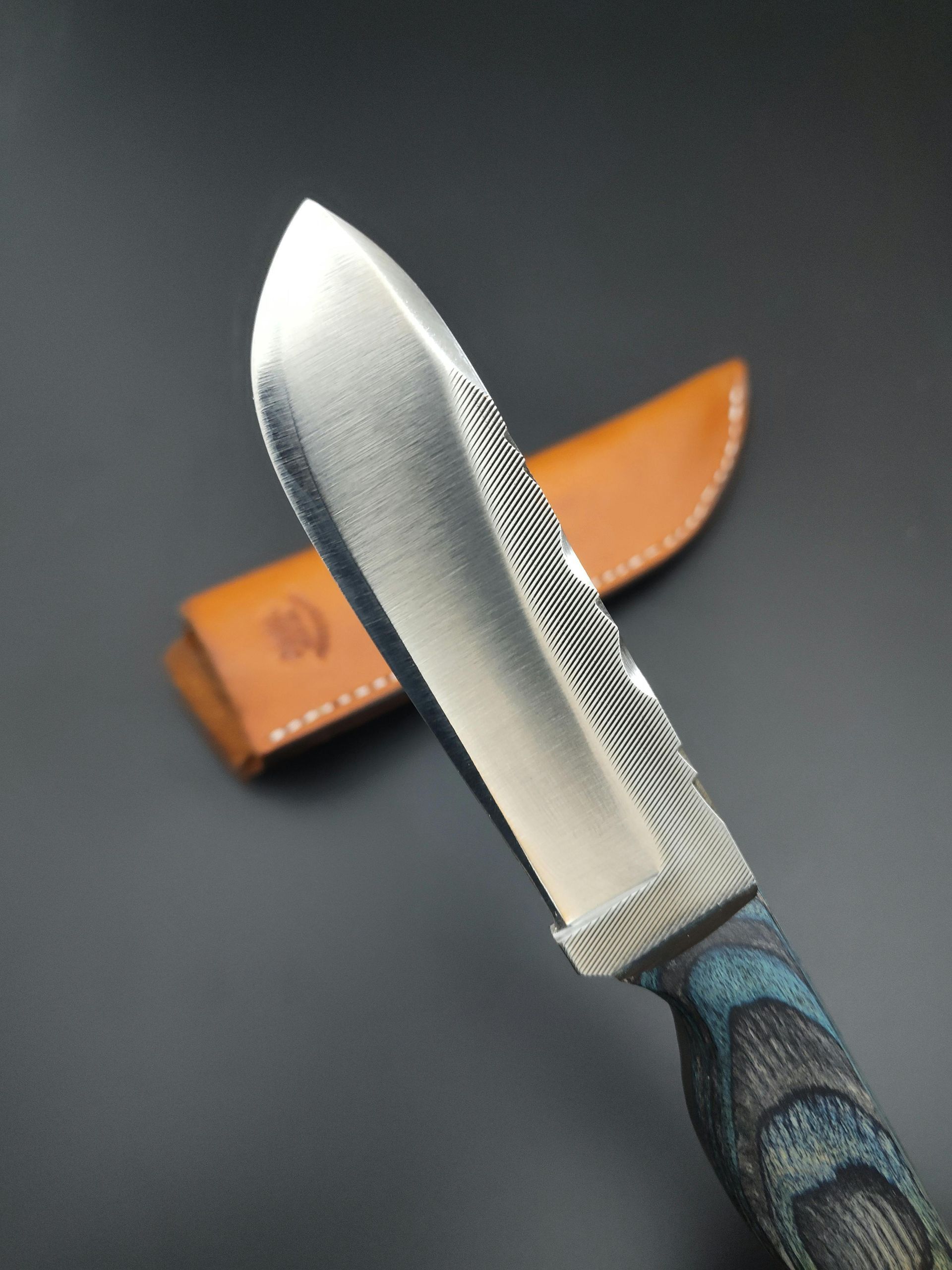 Close-up of a knife with a textured blade and blue-gray handle, set against a leather sheath on a dark surface.