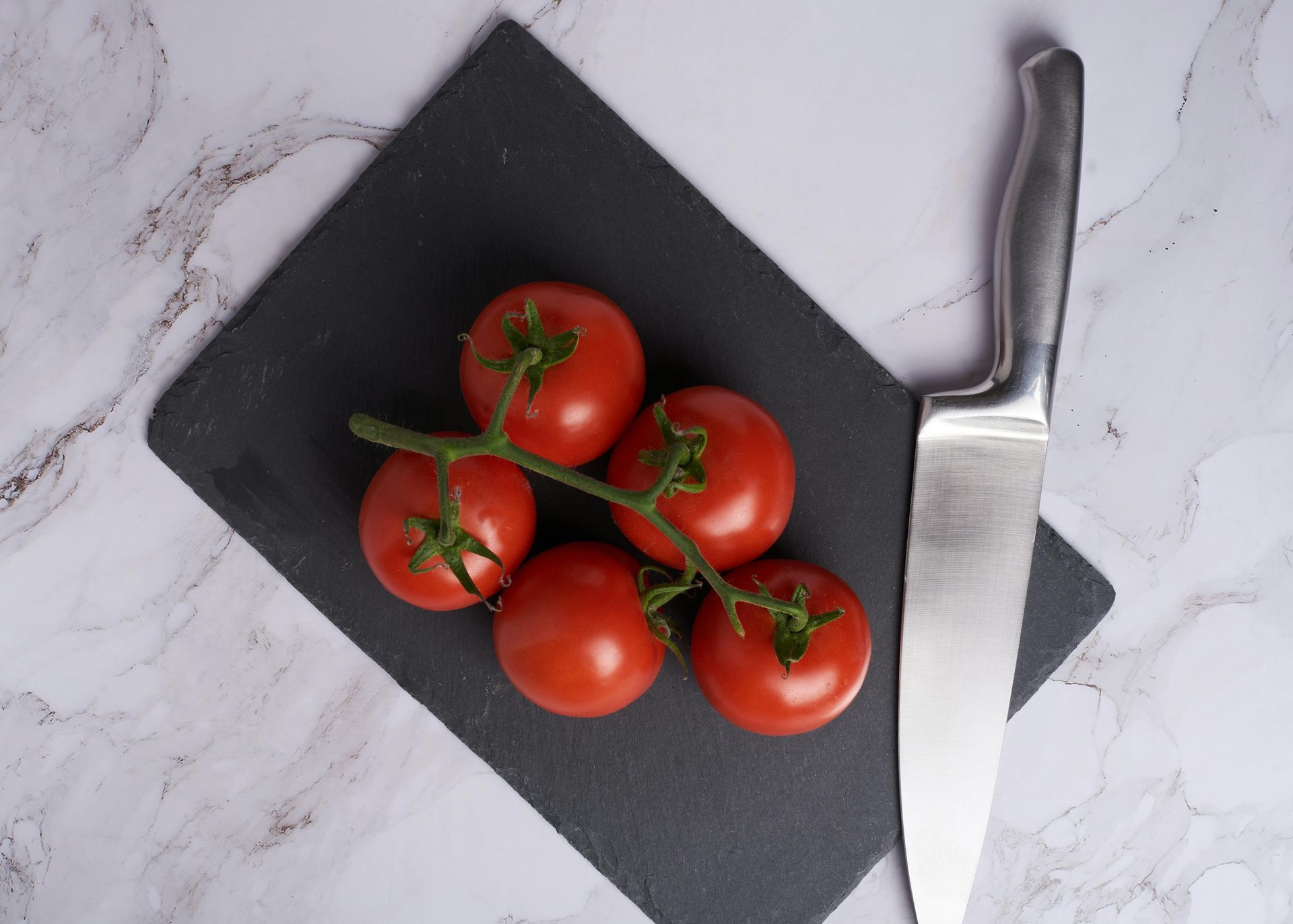 Cherry tomatoes on a black cutting board beside a kitchen knife on a marble surface