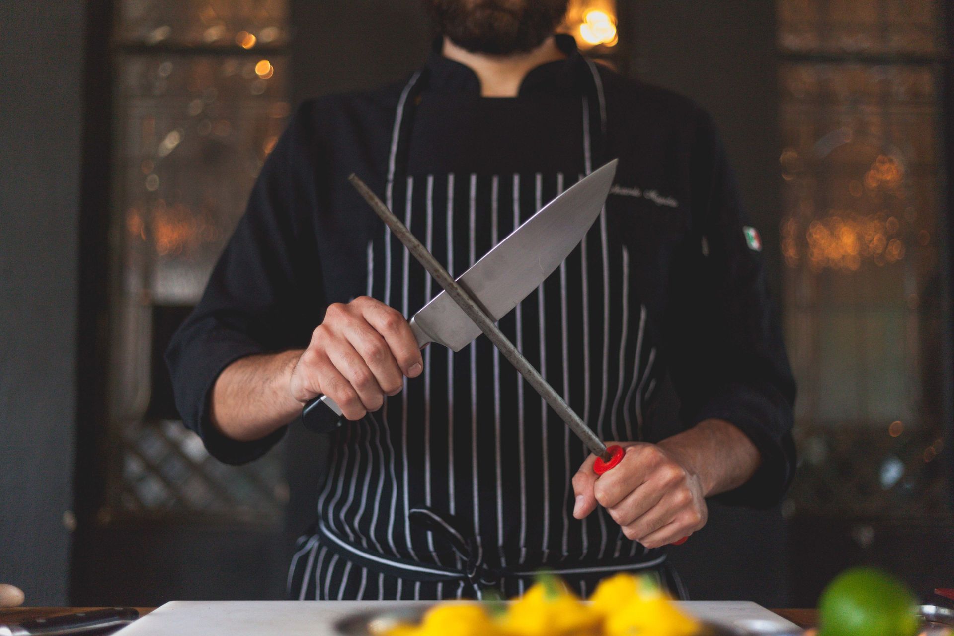Chef sharpening a knife over a striped apron in a kitchen, with fruit on the counter.
