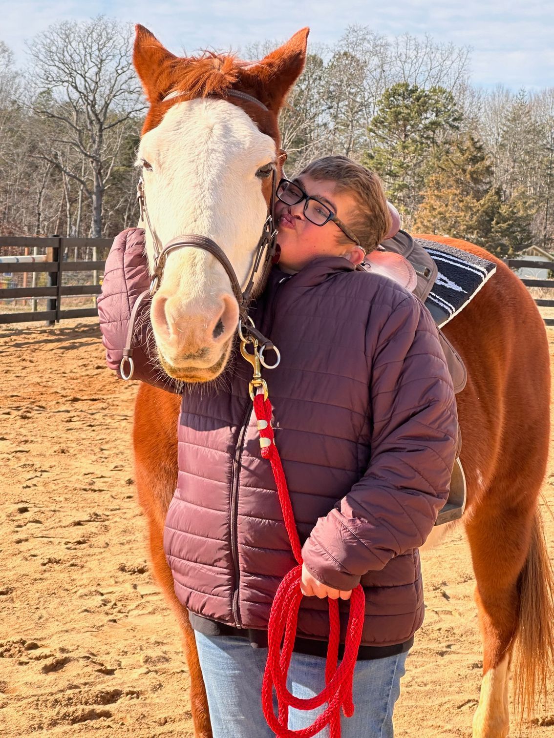 Woman in jacket hugs a chestnut horse with a white face in an outdoor arena.