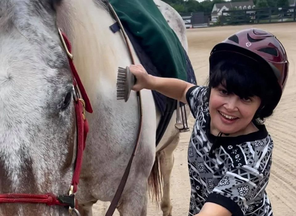 Young person brushes a horse's side in an outdoor riding arena, wearing a helmet and smiling, white horse.