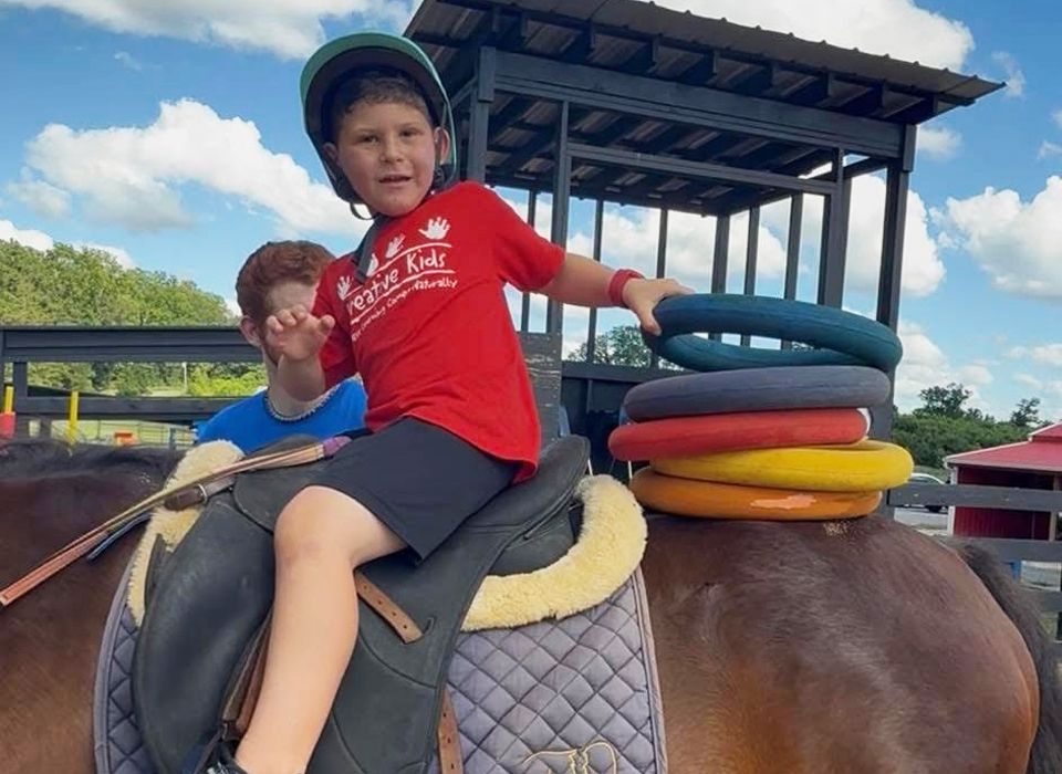 Boy on a horse with rings, wearing helmet, another boy behind him. Outdoors, blue sky.