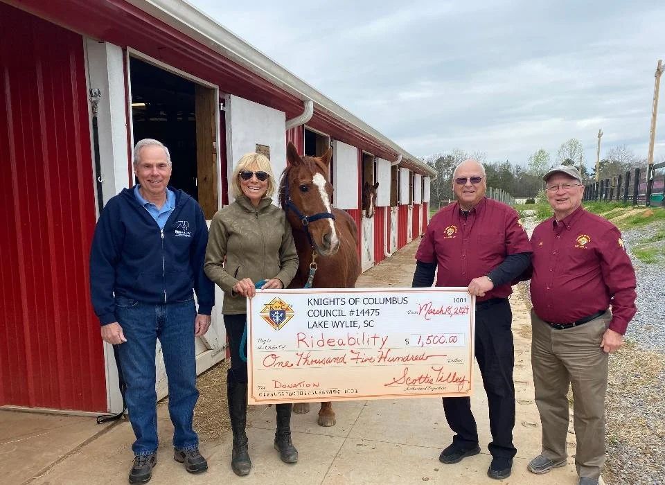 Four people stand with a horse in front of a stable; they hold a large check, smiling.