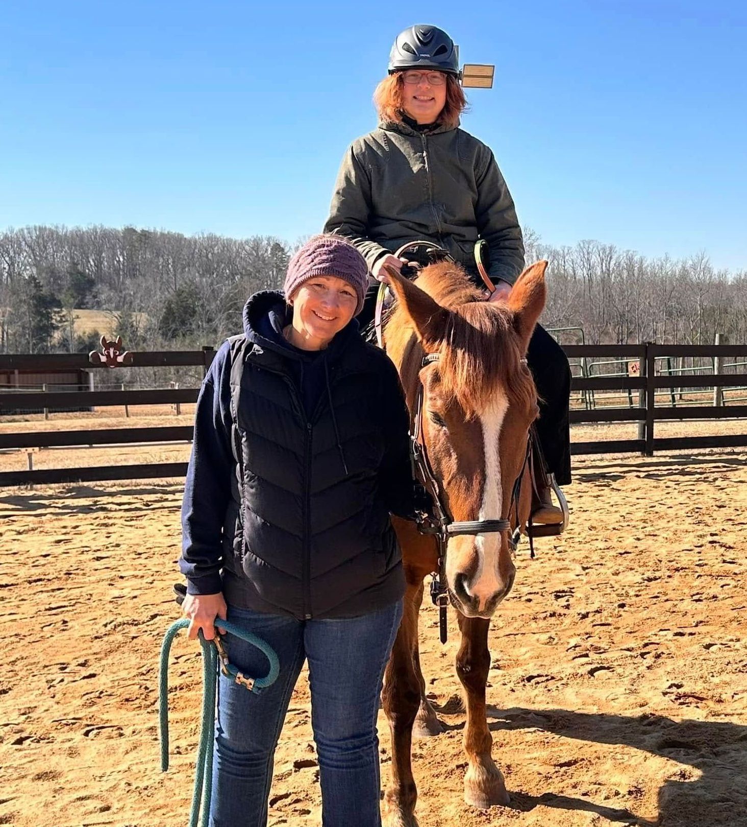 Woman on horse, another beside, smiles. Outdoors, sunny day, riding arena.
