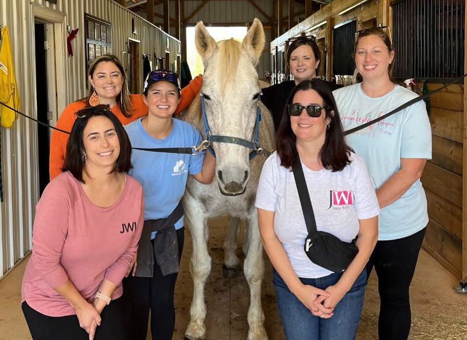 Group of women pose with a light-colored horse in a stable.