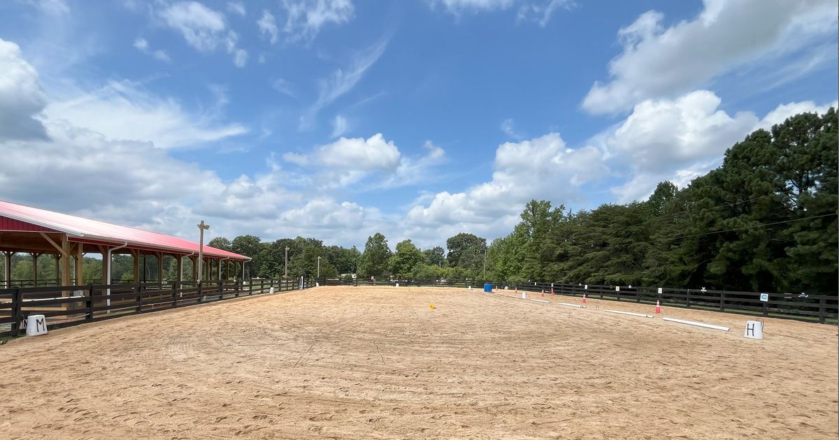 Equestrian arena with tan sand, a red-roofed shelter, and trees against a blue sky with clouds.