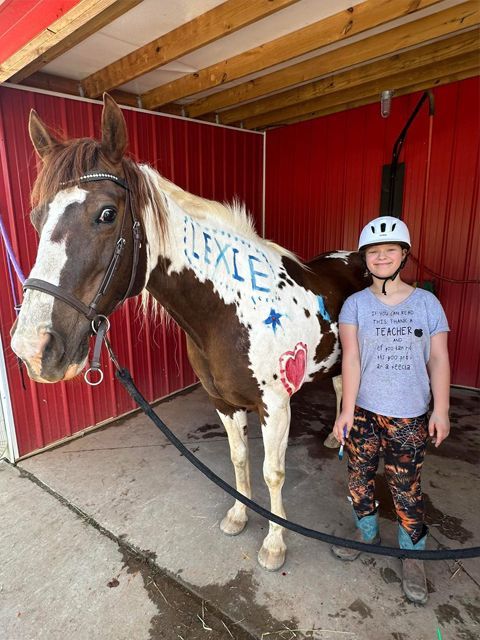 A girl in a riding helmet poses with a horse named Lexie in a red stable.