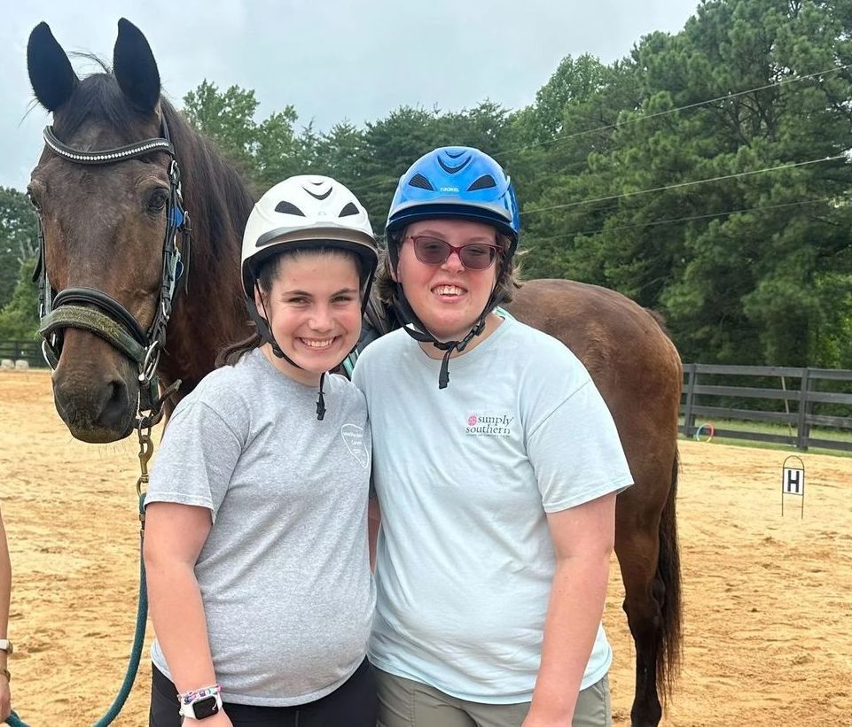 Two smiling people with a horse, all wearing helmets, standing in a sandy arena.