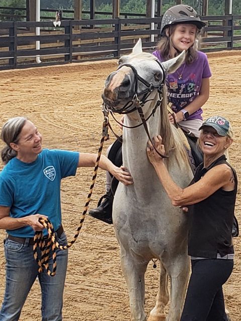 Two women and a girl smile while petting a gray horse in an outdoor riding arena.