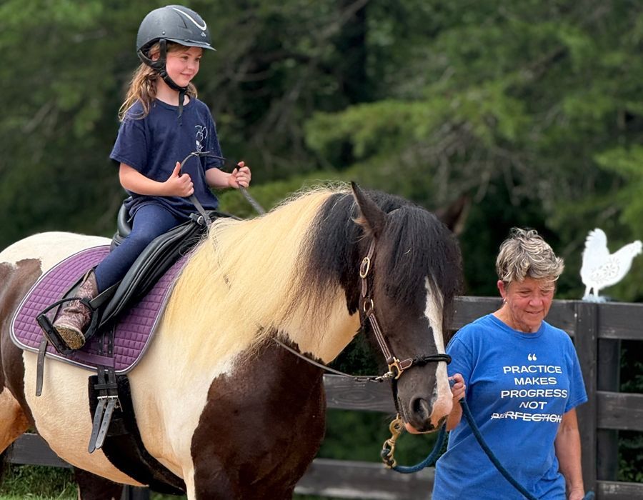 A child on a pony holds an Easter egg while being assisted by a woman at an outdoor riding arena.