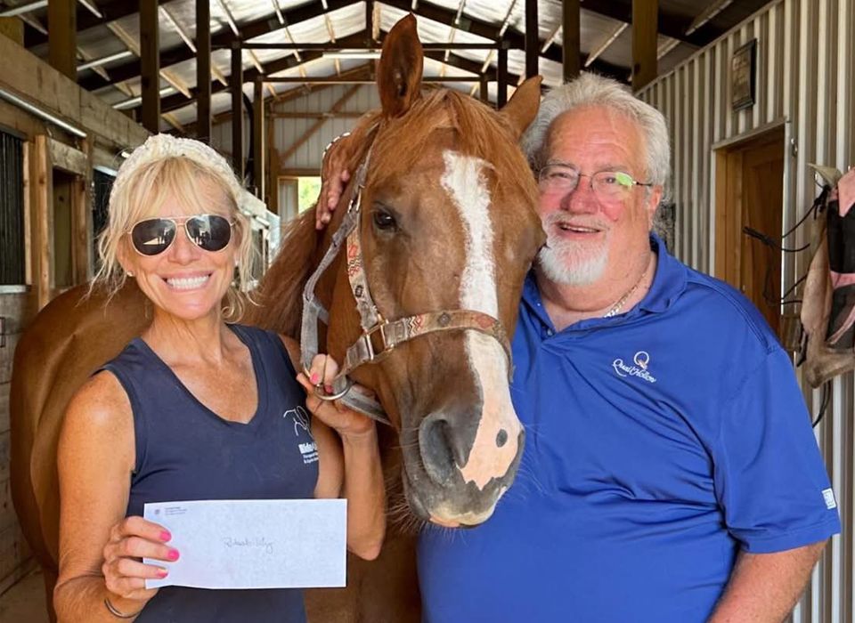 Woman and man with a chestnut horse in a barn. Woman holds a check; both smile.