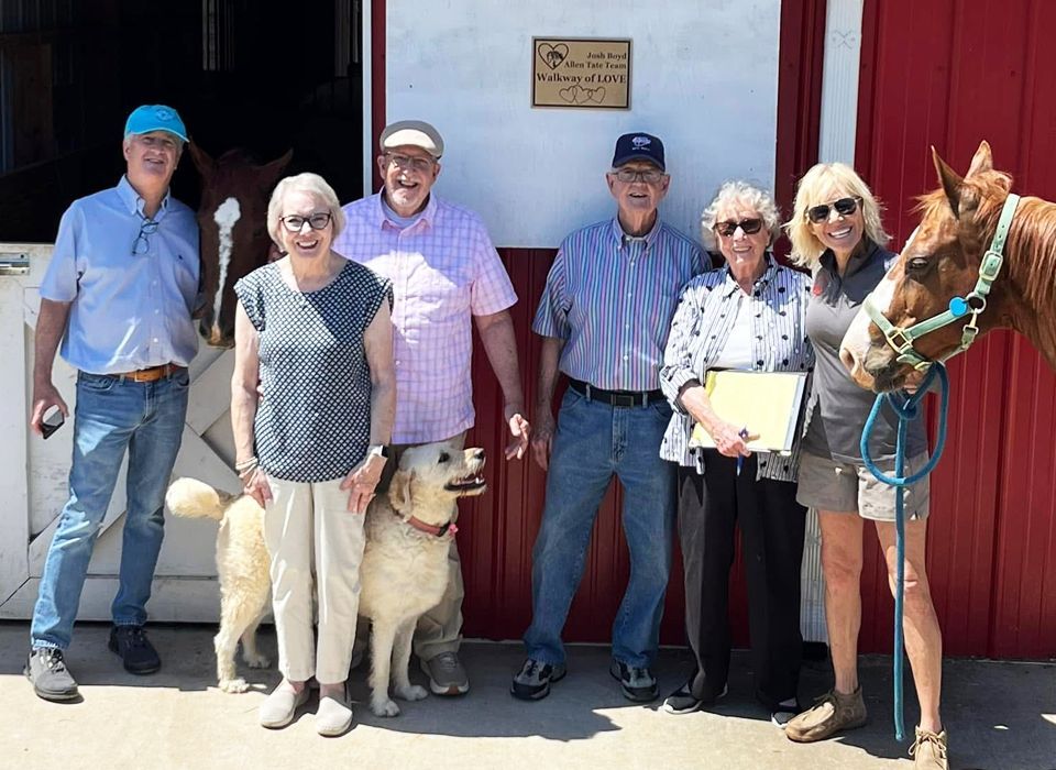 Group poses with horses and a dog outside a red barn.