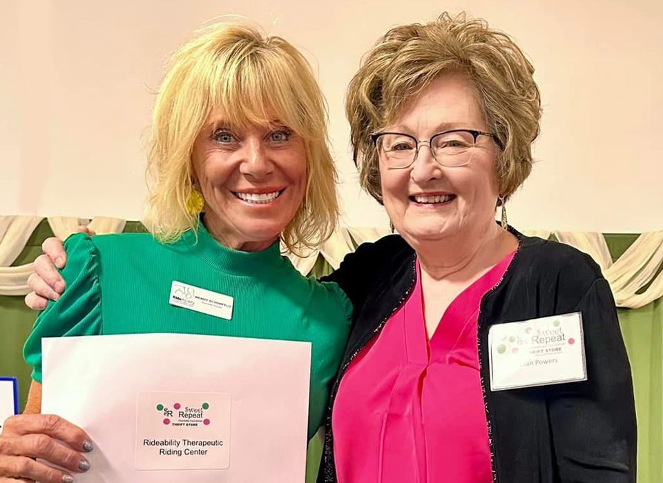 Two women smiling, one holding a paper, in a room with a curtain. The woman on the left wears green, the other pink.
