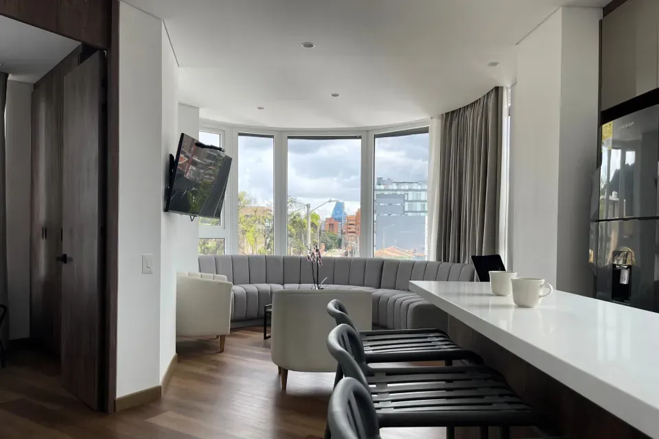 Amplia sala con sofá en L, cocina moderna y ventanales con vista urbana en la Apartasuite Deluxe de VOU Suites Bogotá, hotel de larga estancia en Bogotá ideal para viajes de negocios y turismo.