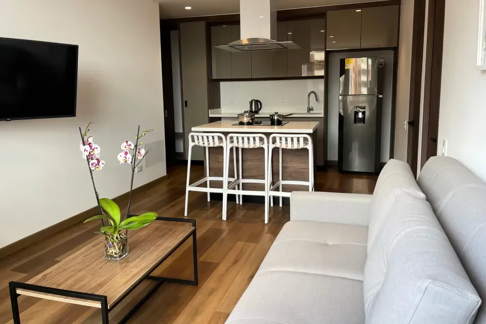 Apartasuite en VOU Suites Bogotá con sala moderna equipada con sofá gris, mesa de centro con orquídea, televisor, cocina integral con nevera, estufa y barra comedor con sillas, ideal para largas estancias en Usaquén Bogotá.