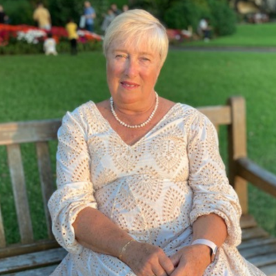 Carol, Tutorio primary school tutor, seated on a park bench wearing a white eyelet dress and pearl necklace