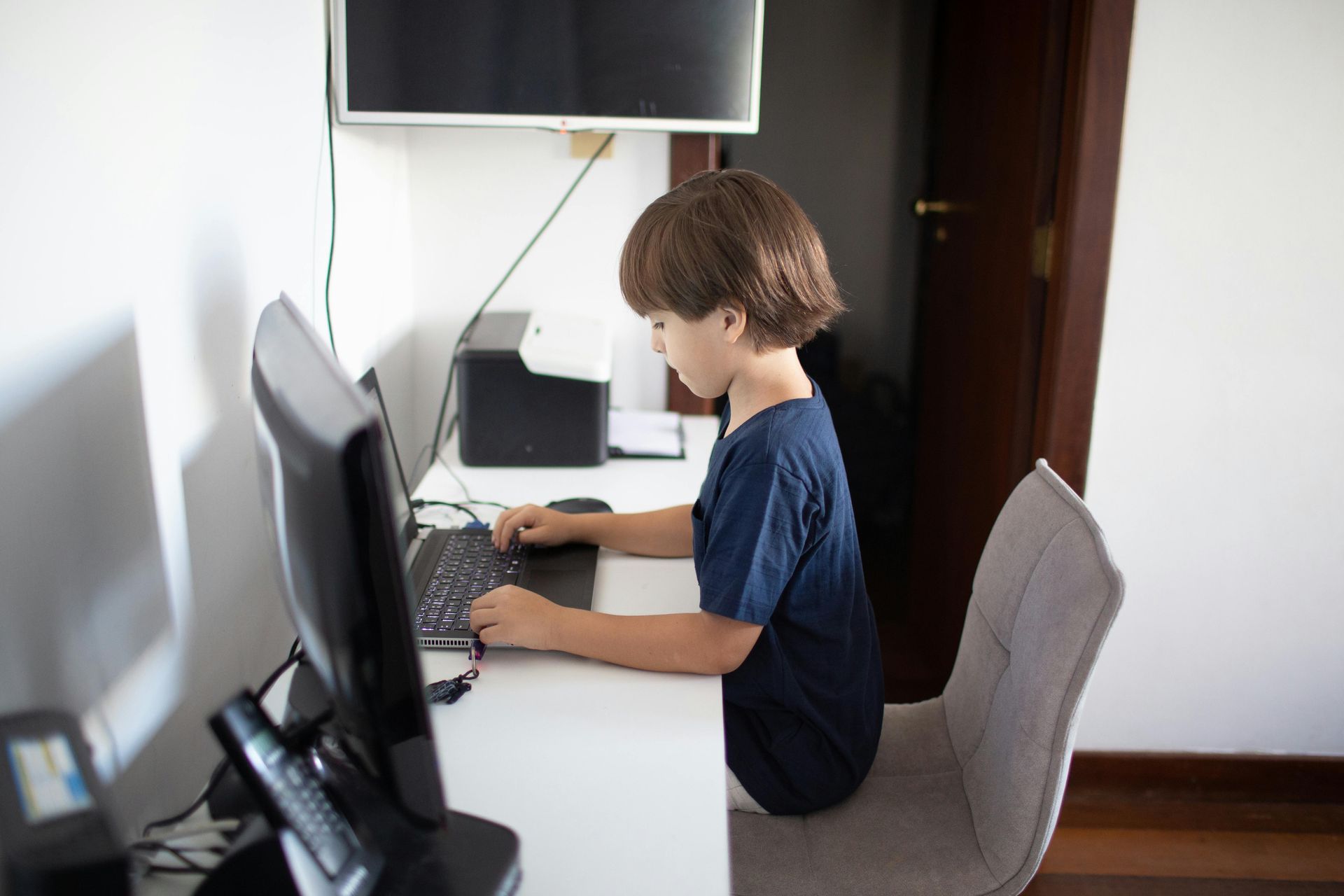 A young boy in a blue shirt types on a laptop at a white desk in a room with a monitor and printer.