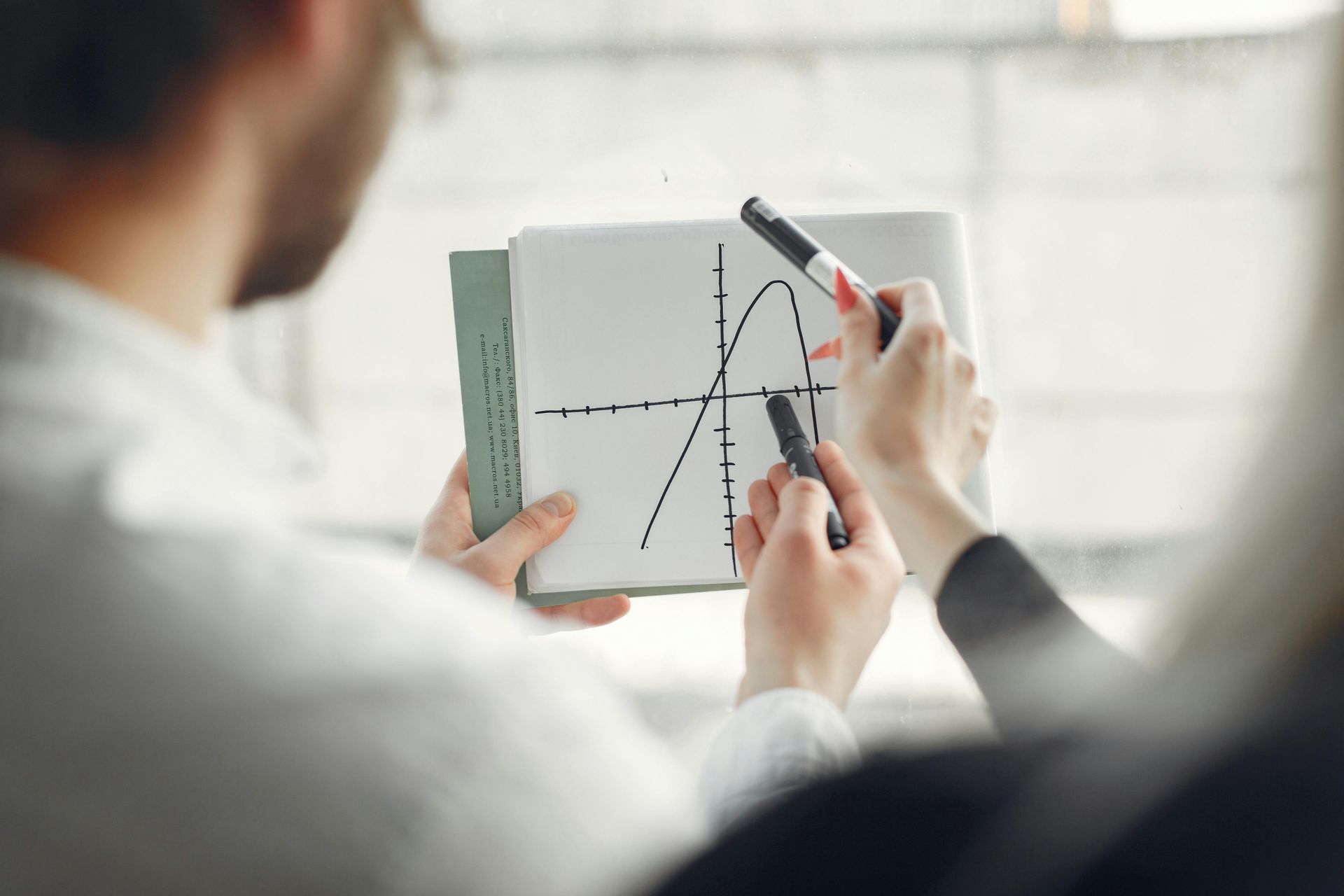 Two people examine a hand-drawn graph of a downward-opening parabola on a notepad during a meeting.