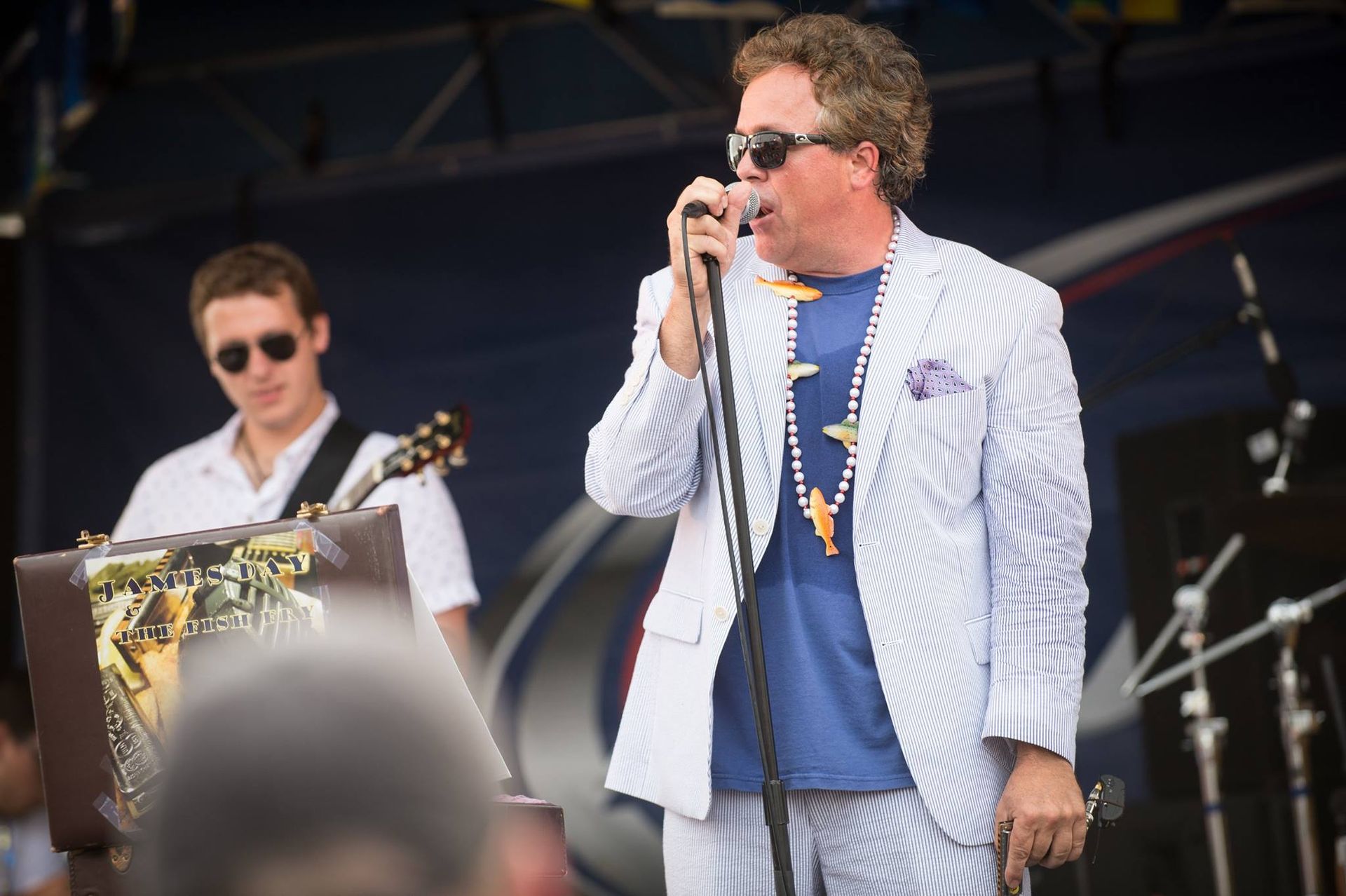 Man in seersucker suit sings into a microphone on stage; guitarist in background.