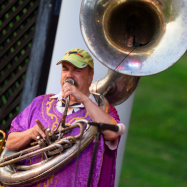 Man playing a tuba and singing into a microphone, wearing a purple shirt and a green cap.