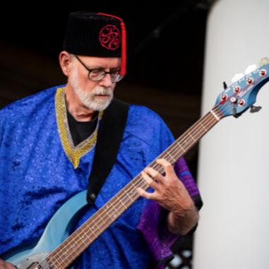 Man playing electric bass, wearing blue robe, black fez with red trim. Performing outdoors.