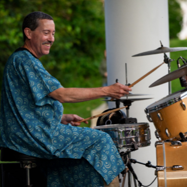 Man playing drums outdoors, wearing a blue patterned shirt.