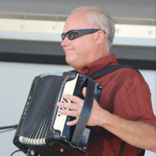 Man in sunglasses playing an accordion outdoors, wearing a red shirt.