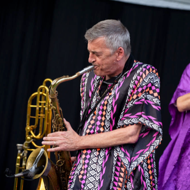 Saxophonist playing a gold saxophone, wearing a colorful patterned poncho, on stage.