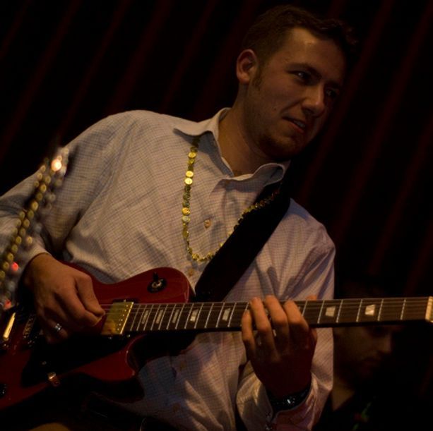 Man playing a red electric guitar onstage, wearing a light blue shirt and gold necklace.