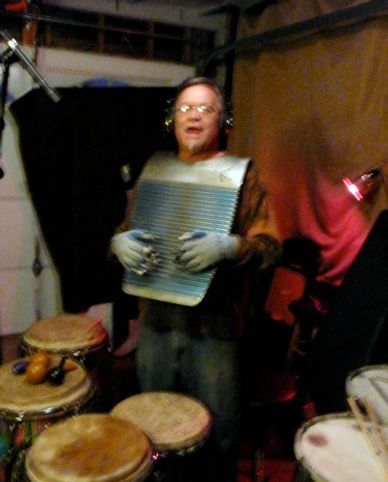 Man playing a washboard as a percussion instrument, wearing gloves and headphones, surrounded by drums.