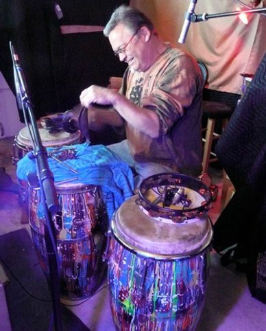 A man seated, playing colorful conga drums, holding a drum, with a tambourine nearby in a dim setting.