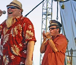 Two men singing on stage. One in red Hawaiian shirt, the other in a striped shirt and cap, both wearing sunglasses.