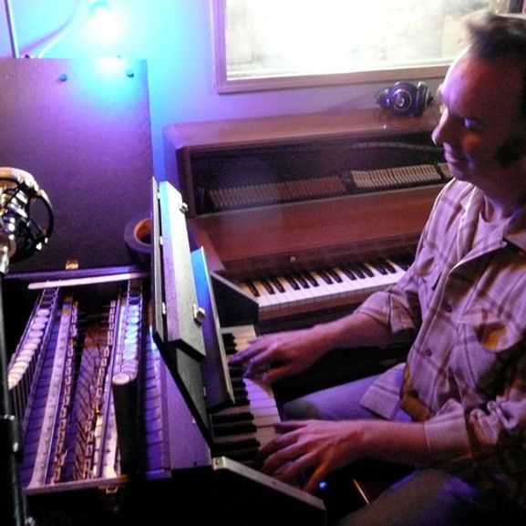 Man playing keyboard in a studio with a microphone and a pipe organ visible.