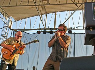Two men performing on stage; one playing guitar, the other harmonica. Outdoors, light colors.