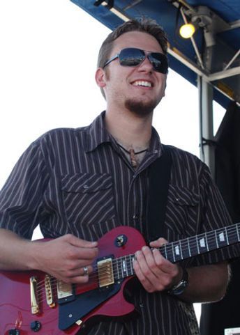 Man playing a red electric guitar on stage, wearing sunglasses and smiling.