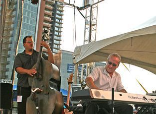Jazz musicians performing outdoors: bassist on left, keyboardist on right, under a tent. City buildings in the background.