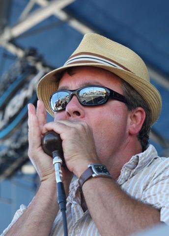 Man playing harmonica into a microphone, wearing a fedora and sunglasses.