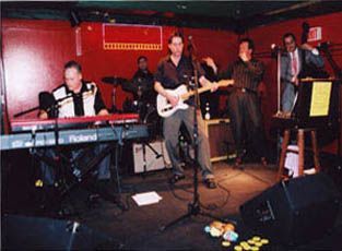 Band performing on stage with red background; keyboard, guitar, bass, drums, and saxophone players.