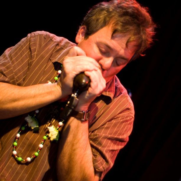 Man playing harmonica, brown striped shirt, beaded necklace, dark stage.