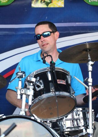 Drummer in sunglasses playing a black drum set, wearing a blue shirt. Outdoors, with a blue background.