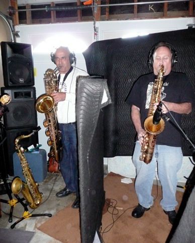 Two men playing saxophones in a recording studio setting, with multiple instruments and audio equipment.