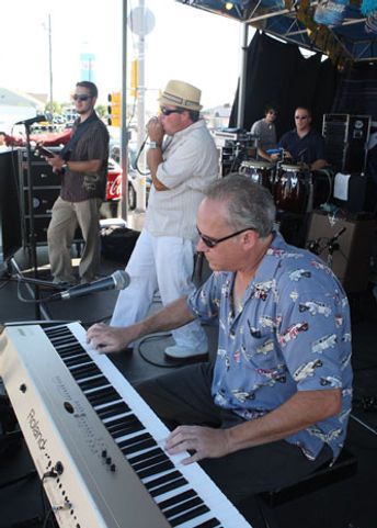 Band performing outdoors, keyboardist in foreground, singer with harmonica, guitarist, drummer in background.
