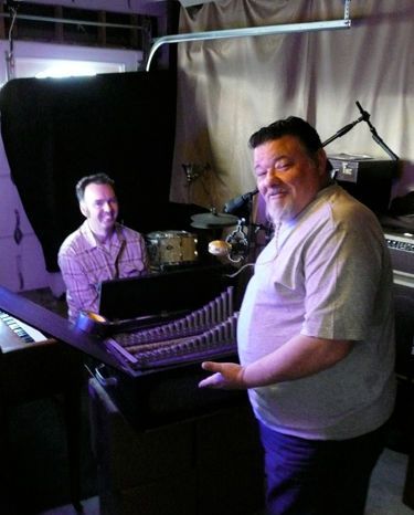 Two men in a dimly lit room, one holding an instrument, another smiling at a keyboard.
