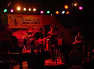 Band performing on a stage lit by colorful lights; Diamond State Blues Society banner.