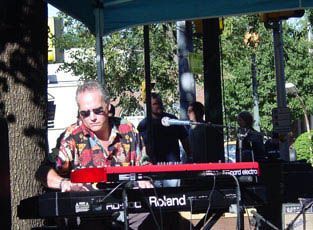 Keyboardist in sunglasses plays a red Nord Electro keyboard outdoors. Other band members and trees in the background.
