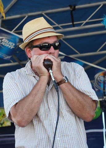 Man playing harmonica on stage, wearing a fedora and sunglasses.