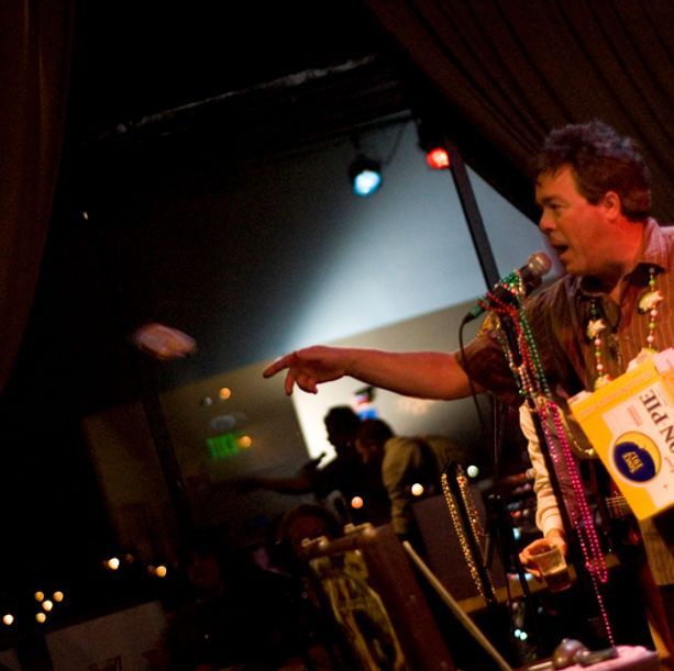 Man onstage throws something, pointing with his right arm. Stage lighting and musicians in background.